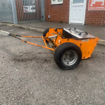Orange compacting machine on a paved lot beside brick buildings and a metal gate, with a white door nearby.