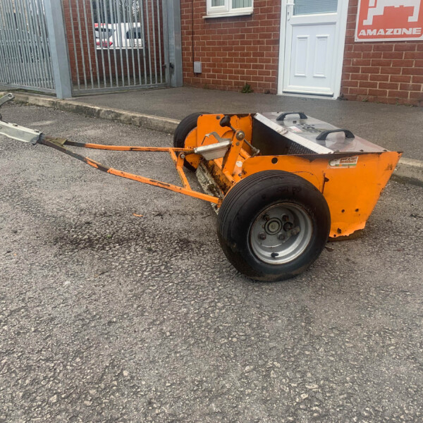 Orange compacting machine on a paved lot beside brick buildings and a metal gate, with a white door nearby.