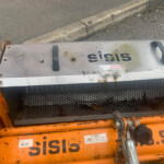 Close-up of an orange industrial machine with a worn metal cover, handles, and a perforated guard, outdoors by a curb.