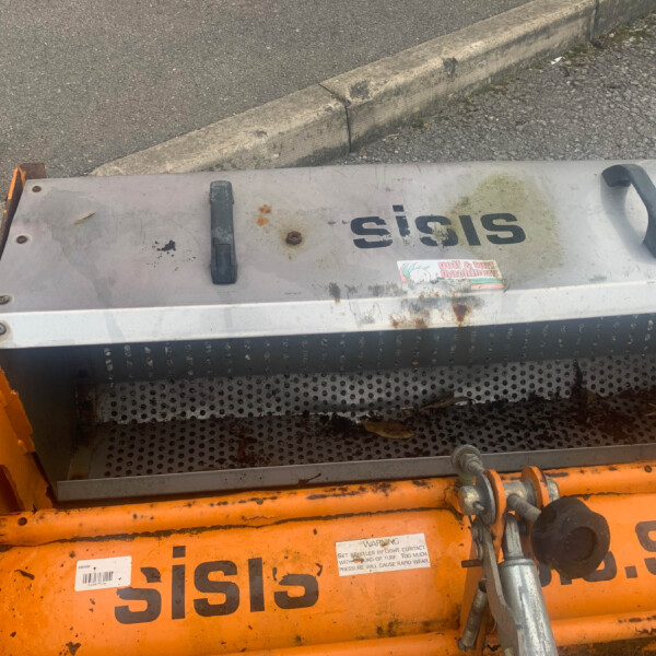 Close-up of an orange industrial machine with a worn metal cover, handles, and a perforated guard, outdoors by a curb.