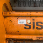Close-up of orange industrial equipment with a barcode label, dirt, and a coin resting on the surface
