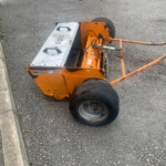 Orange road roller parked on asphalt beside a curb, with large black wheels and a metal frame.