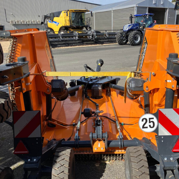 Front view of orange heavy machinery with large tires and hydraulic arms in a yard; a yellow combine harvester and a blue tractor sit in the background beside a metal building.