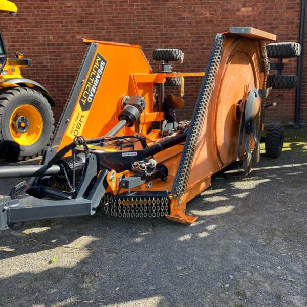Orange industrial equipment with chain-covered components and a conveyor, parked against a red brick wall; a yellow vehicle is partially visible on the left.