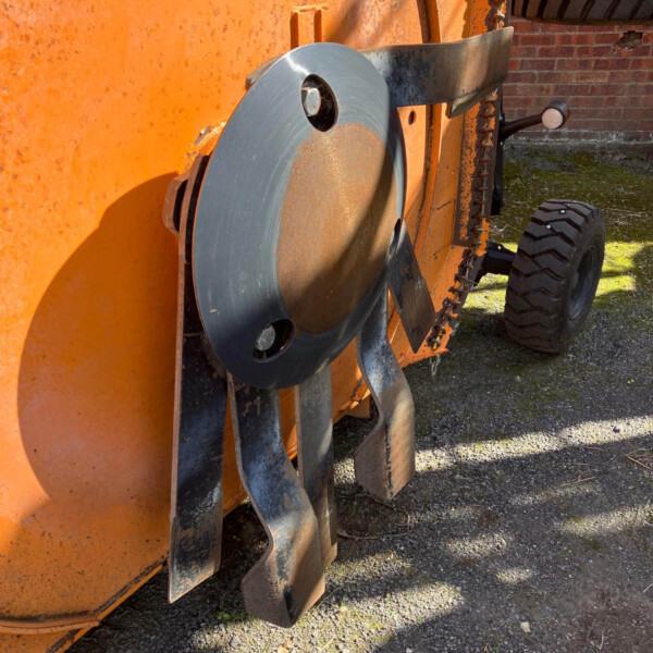 Close-up of an orange industrial machine showing a large circular metal disc and metal guards, with a rugged tire and chain in view on a gravel surface