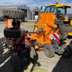 Orange heavy machinery with large tires in an outdoor equipment yard, with grey tents in the background.