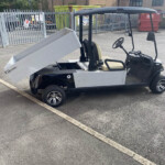 Utility golf-buggy-style vehicle with a raised aluminum cargo bed in a fenced industrial yard.