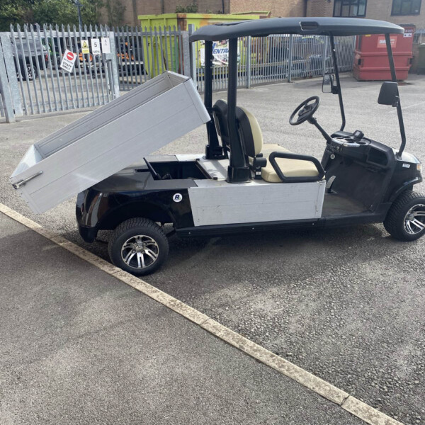 Utility golf-buggy-style vehicle with a raised aluminum cargo bed in a fenced industrial yard.