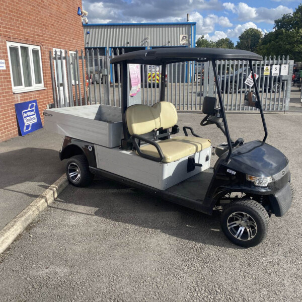 A small utility golf cart with beige seats and an open cargo bed parked beside a brick building outside an industrial gate.