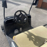Interior view of a golf cart cockpit: steering wheel, pedals labeled STOP on the floor, tan seat, and side mirrors; outdoor pavement and a brick building in the background.