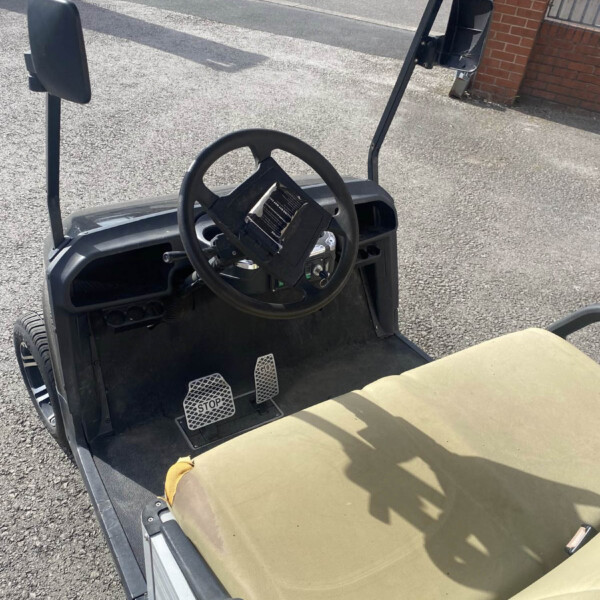 Interior view of a golf cart cockpit: steering wheel, pedals labeled STOP on the floor, tan seat, and side mirrors; outdoor pavement and a brick building in the background.