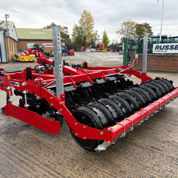 Red heavy‑duty farming implement with black tines in the foreground at an industrial equipment yard; Russell's signage visible in the background