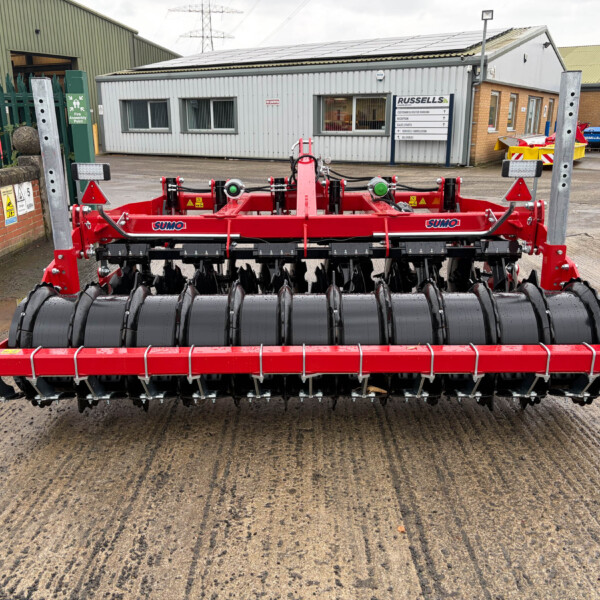 Red agricultural implement with multiple black rolling drums in a farmyard, labeled SUMO, likely a cultivator or seed drill ready for use.