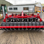 Red agricultural cultivator with black roller discs waiting in a yard outside a workshop building.