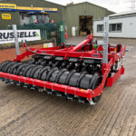 Red agricultural roller cultivator with large black rollers, parked outside a farm machinery workshop.