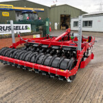 Red agricultural roller implement with black drums sits in a parking area outside a farm workshop row, Russell's sign visible in the background under a green metal building.