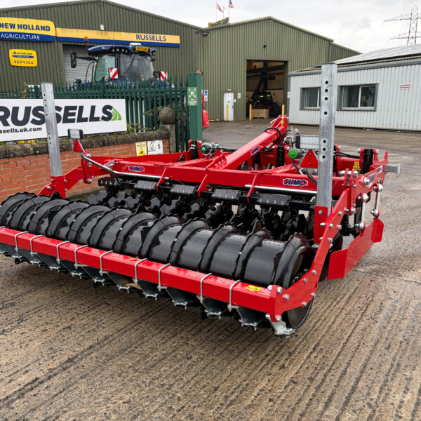 Red agricultural roller implement with black drums sits in a parking area outside a farm workshop row, Russell's sign visible in the background under a green metal building.