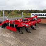 Red agricultural cultivator with blades in a parking area; a Russell's sign is visible in the background.
