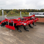 Red Sum o-brand agricultural cultivator with blades and hydraulic hoses, parked on a paved surface near a brick wall and a Russell's sign with autumn trees in the background.