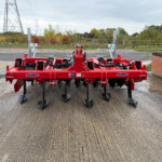Red multi-section farm cultivator with tines on a concrete pad, rural backdrop of trees and power lines.