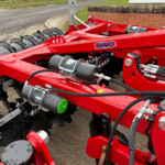 Close-up of a red SUMO agricultural implement with hydraulic hoses, metal plates, and a green cap, showing its machinery components.