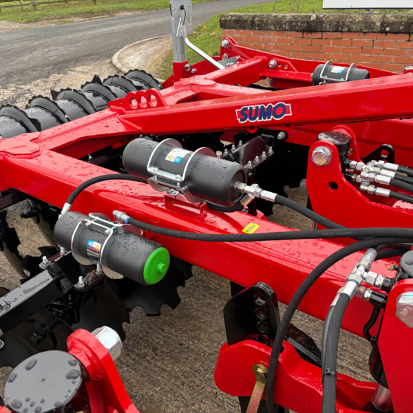 Close-up of a red SUMO agricultural implement with hydraulic hoses, metal plates, and a green cap, showing its machinery components.