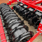 Close-up of a red agricultural cultivator with overlapping black disc blades attached to a metal frame, wet from rain.