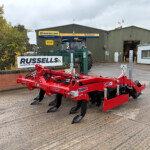 Red agricultural machinery on a paved yard in front of a green warehouse with Russell's signage and a hedge nearby.