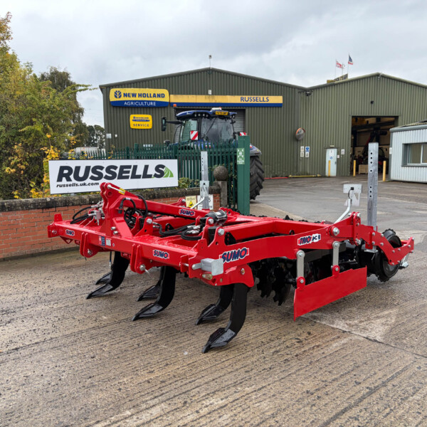 Red agricultural machinery on a paved yard in front of a green warehouse with Russell's signage and a hedge nearby.