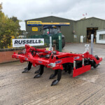 Red agricultural implement with black tines parked on a concrete lot outside a green metal warehouse with Russell's sign in view.