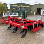 Red agricultural implement with multiple tines in the foreground at a farm service yard, Russell's building and tractor visible in the background.
