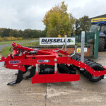 Red agricultural equipment in the foreground with a blue New Holland tractor and 'RUSSELLS' sign in the background.