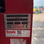 Close-up of a red machine with a metal nameplate reading 'TOMAHAWK 8500', showing 'Serial No' and model/year details; farm equipment visible in the background.