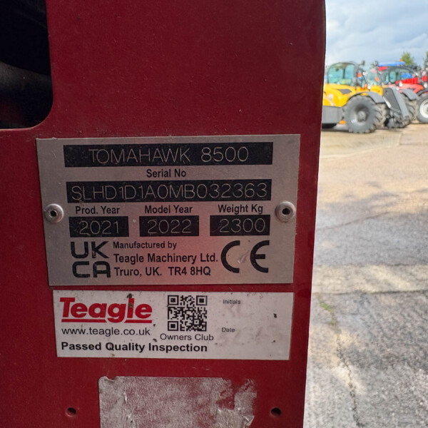 Close-up of a red machine with a metal nameplate reading 'TOMAHAWK 8500', showing 'Serial No' and model/year details; farm equipment visible in the background.