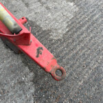 Red hydraulic jack with a yellow support bar on a gravel surface, close-up detail of the tool.