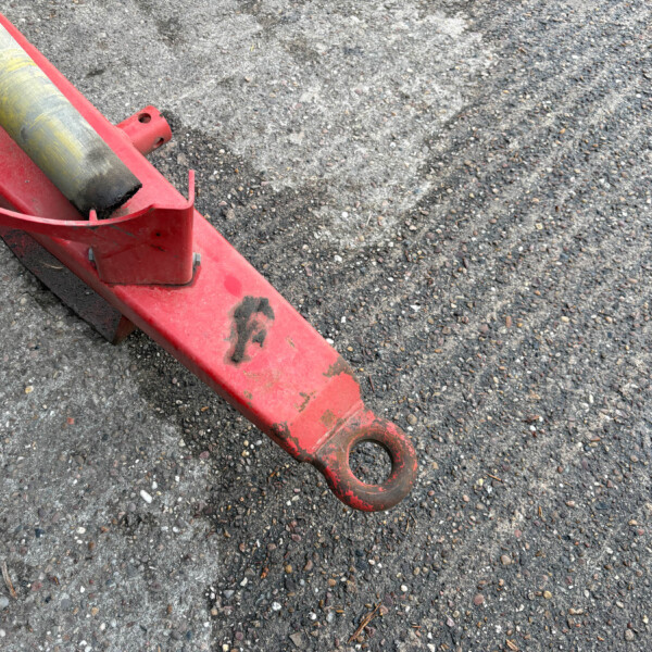 Red hydraulic jack with a yellow support bar on a gravel surface, close-up detail of the tool.