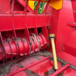 Close-up of a red harvesting header with rotating sickle blades cutting stalks; a yellow PTO drive shaft runs along the side.