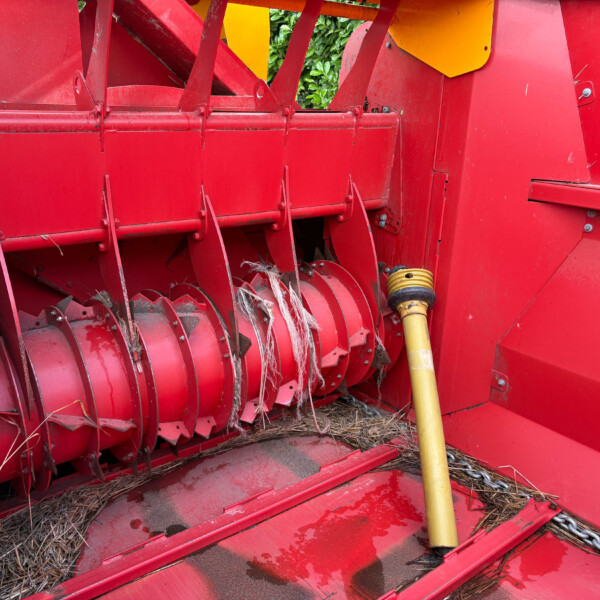 Close-up of a red harvesting header with rotating sickle blades cutting stalks; a yellow PTO drive shaft runs along the side.