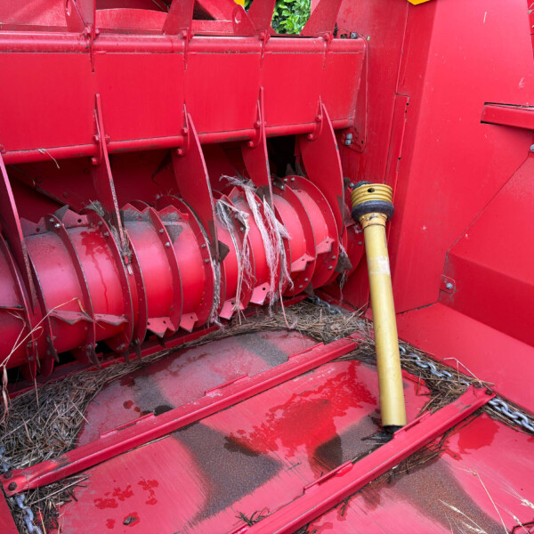 Close-up of a red agricultural harvester header with rotating blades and tangled straw, plus a yellow PTO shaft nearby