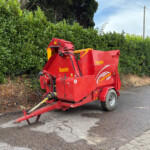 Red Teagle Tomahawk 8500 bale baler parked beside a dense green hedge on a rural road.