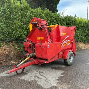 Red Teagle Tomahawk 8500 forage harvester parked beside a tall green hedge on an asphalt road.