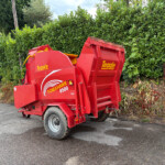 Red Teagle Tomahawk 8500 bale processing machine in a paved lot beside a tall green hedge, facing left.