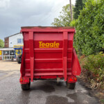 Rear view of a red Teagle trailer with soil on top, parked in a dealership lot beside a tall green hedge.