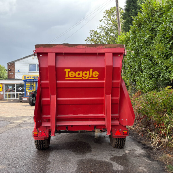 Rear view of a red Teagle trailer with soil on top, parked in a dealership lot beside a tall green hedge.