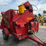 Red Teagle hay baler with yellow chute and hydraulic hoses, parked in a lot with other yellow farm equipment visible in the background.