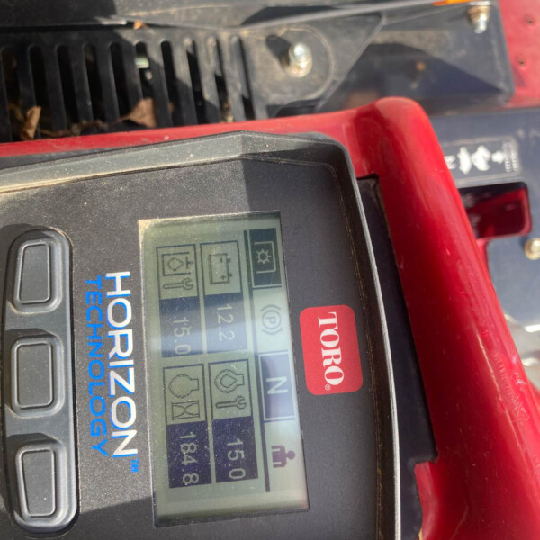 Handheld diagnostic meter with a touchscreen showing icons and a green status light on a red surface.