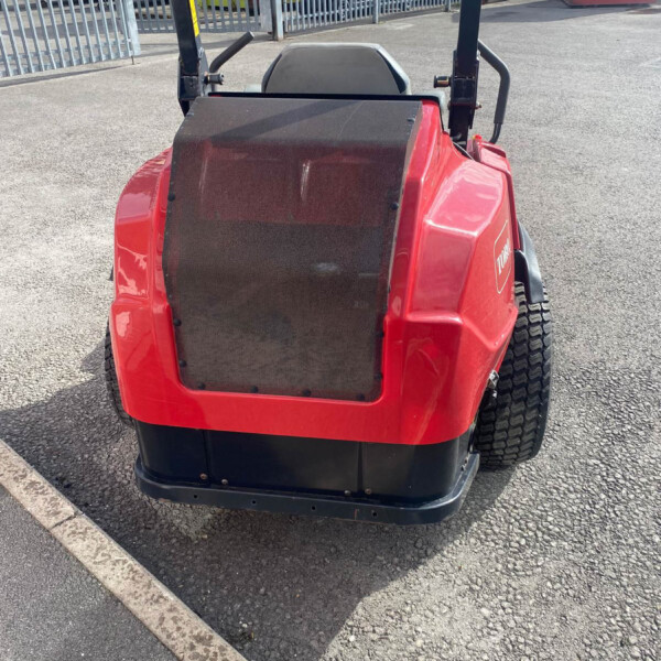 Red ride-on lawn mower photographed from the rear in a parking lot, showing seat, roll bar, and large rear wheels near a curb and metal fence.