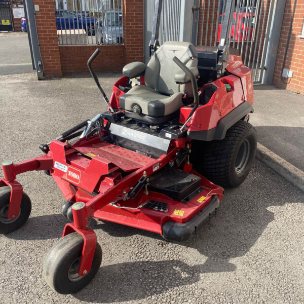 Red Toro Z Master riding lawn mower with gray seat, parked in a paved area near a tall metal gate.