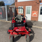 Red Z-Master ride-on lawn mower parked in a parking area in front of a brick building with Russell's sign and an Amazon logo sign nearby.