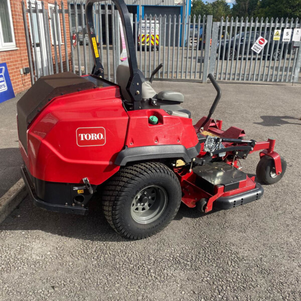 Red Toro riding lawn mower parked in an outdoor lot beside a brick building and metal fence, with a mower deck and seat visible.